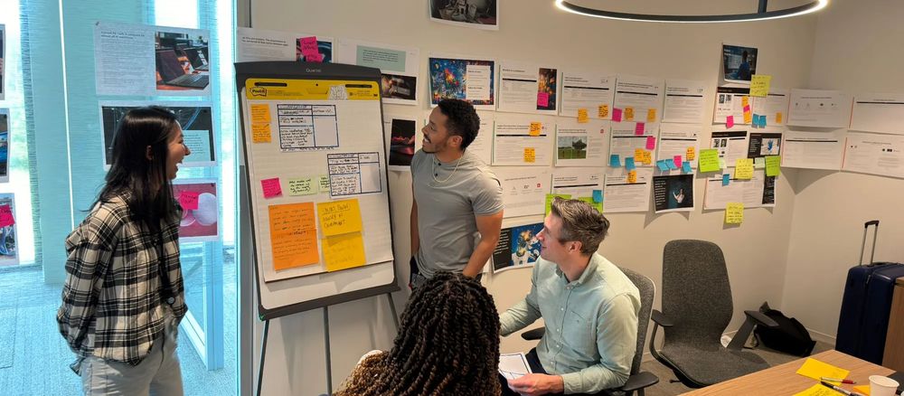 Four people around a flipchart in a workshop room, with a wall of printed customer research and sticky notes behind them